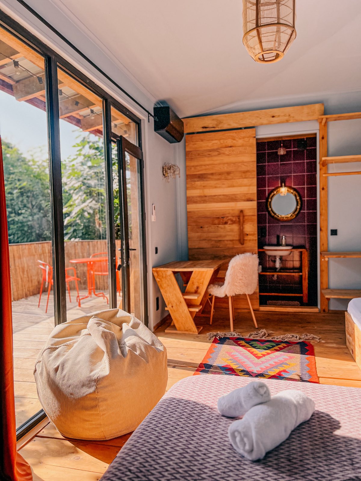 A bright cabin room with sliding glass doors, a wooden desk and an en-suite bathroom with plum-coloured tiles.