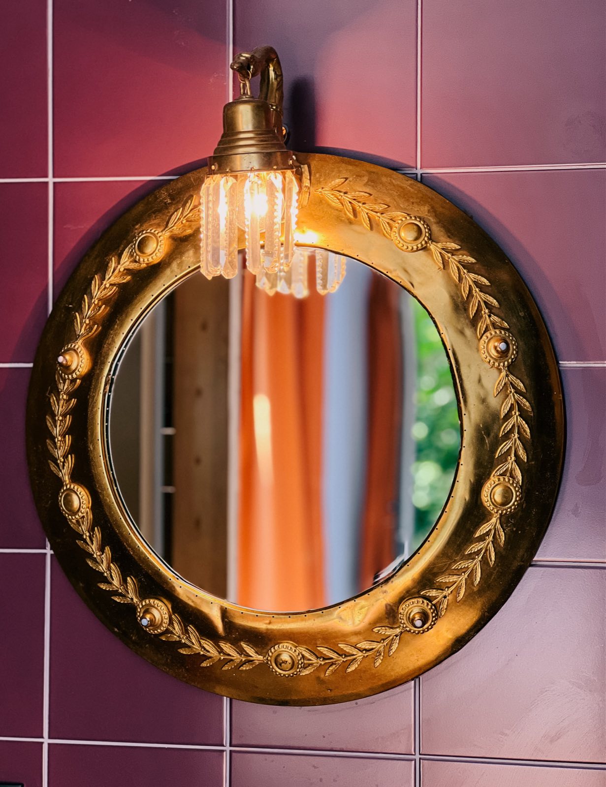 A round brass mirror with a vintage crystal lamp on plum-coloured bathroom tiles.