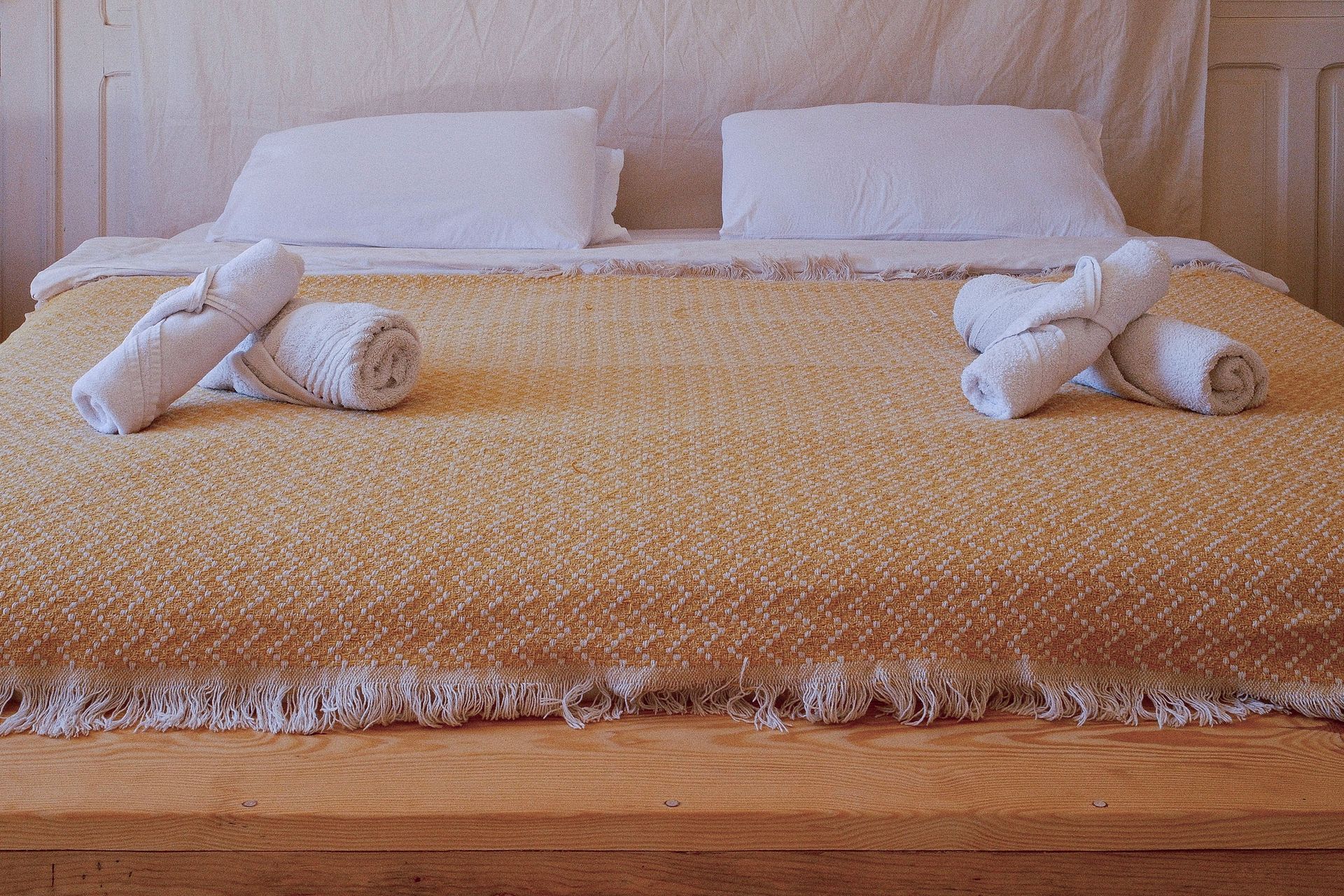 A neatly made bed with white pillows, a textured yellow fringed blanket, and two sets of rolled white towels.