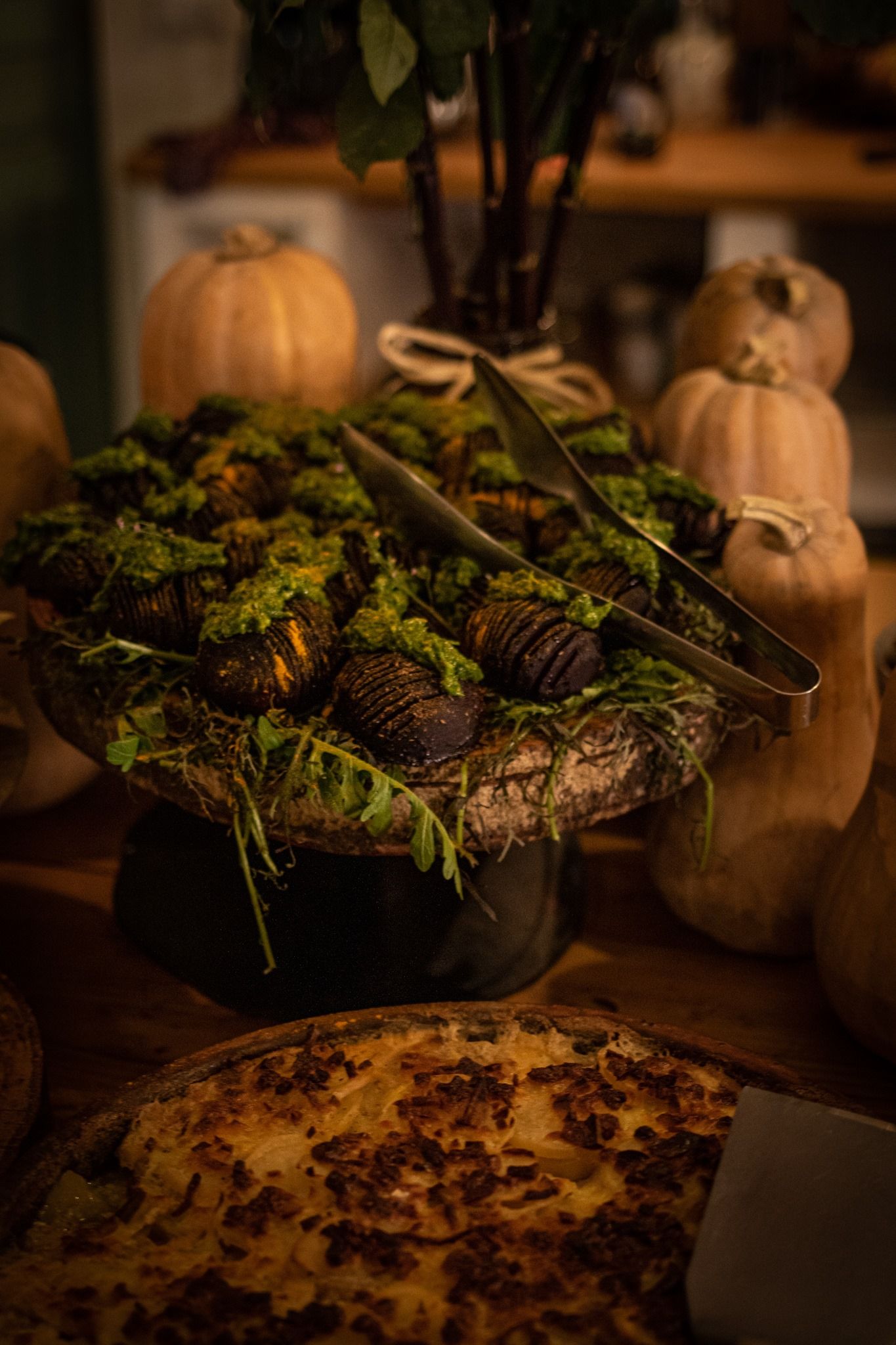 A rustic buffet table features baked goods on a pedestal tray, served with tongs and surrounded by decorative pumpkins.