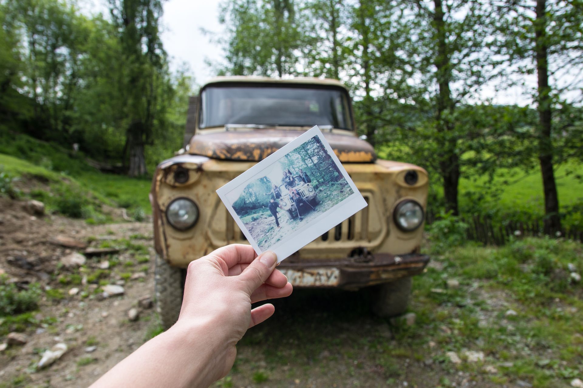 A hand holds a photograph against a weathered, beige truck parked in a lush, green, wooded setting.