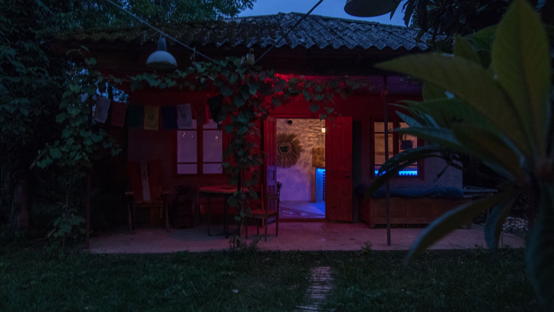 A small cottage at dusk with a red-lit exterior and an open doorway revealing a brightly lit, blue-hued interior.
