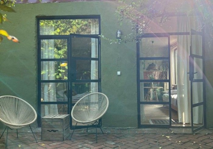 Two light-colored Acapulco-style chairs sit on a brick patio in front of a green wall with two glass-paned French doors.