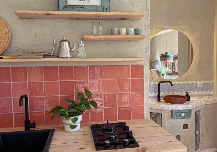 A rustic kitchen with a wooden counter, pink tiled backsplash, black sink and stove, and open shelving against a wall.