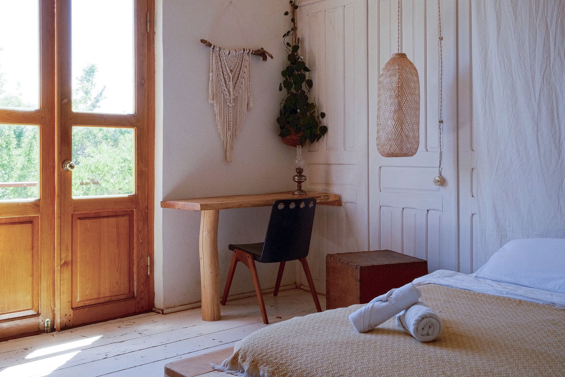 A rustic bedroom with a wooden desk, a dark chair, a macramé wall hanging, a hanging lamp, and a bed with rolled towels.