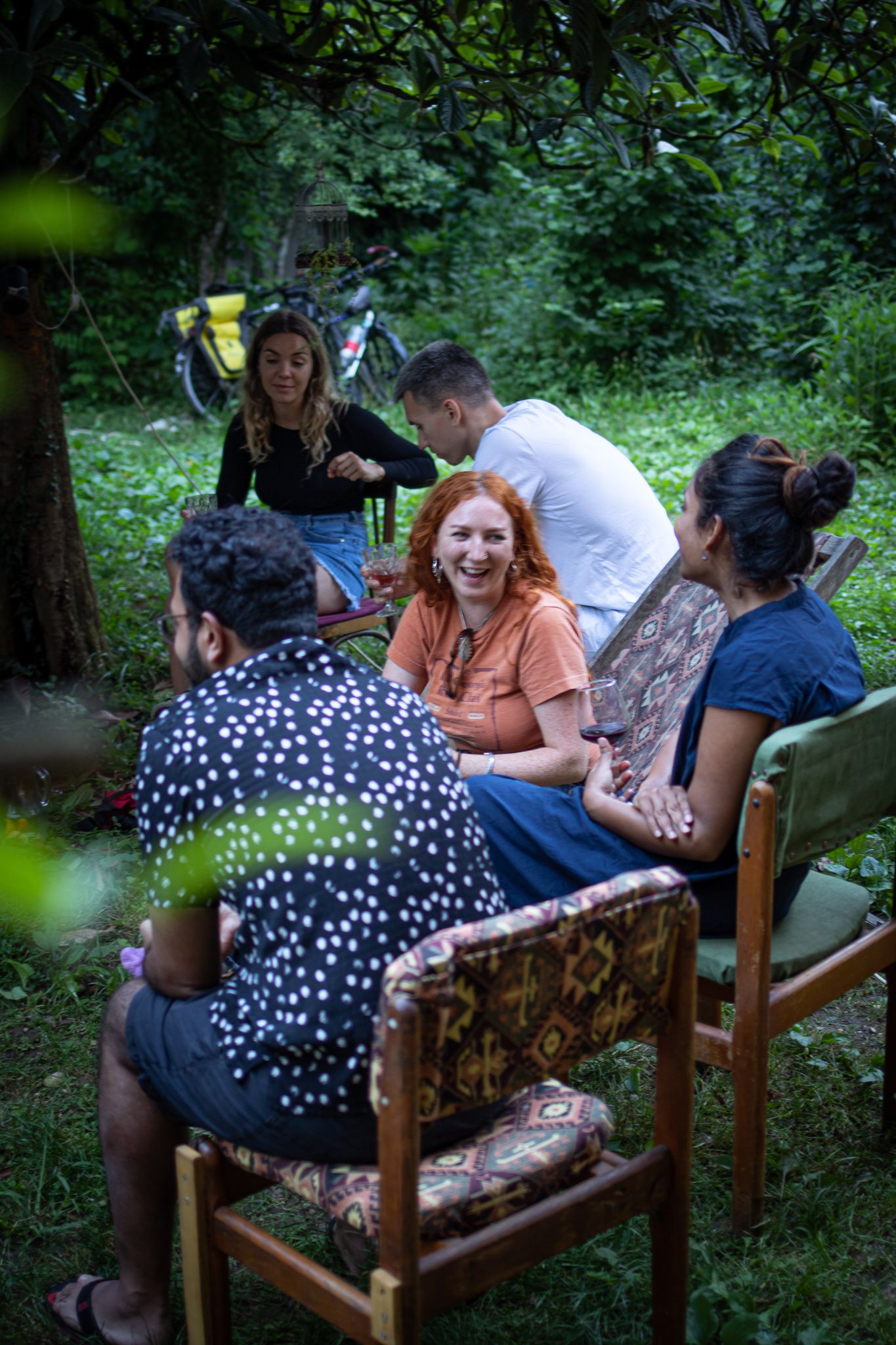 A diverse group of people sitting in a garden, laughing and talking in vintage chairs.
