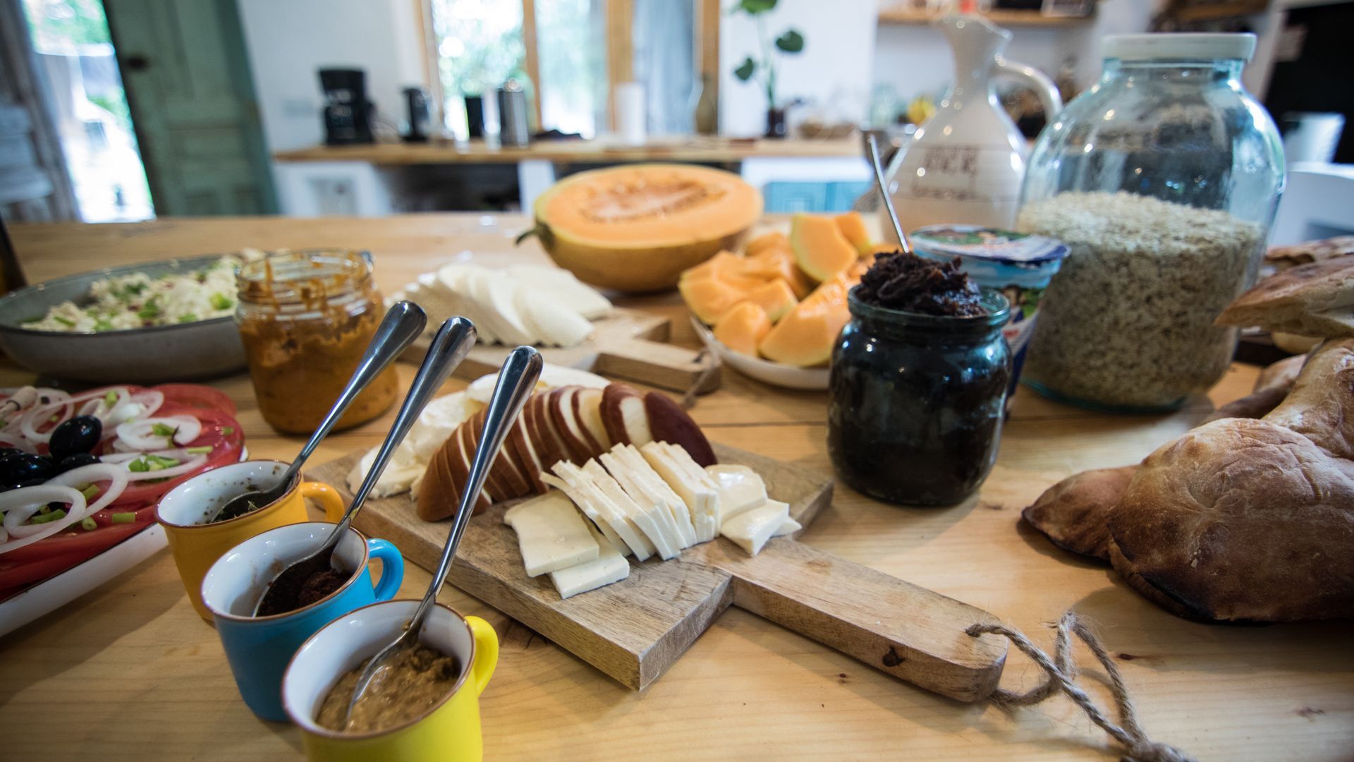 A rustic wooden table is spread with a breakfast buffet featuring sliced bread, cheese, melon, spreads, and grains.