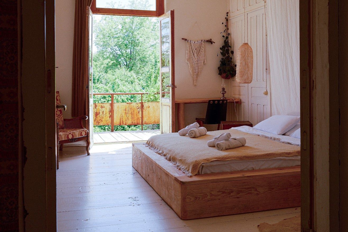A sunlit bedroom featuring a minimalist wooden bed frame, a balcony door opening to trees, and boho-style wall decor.