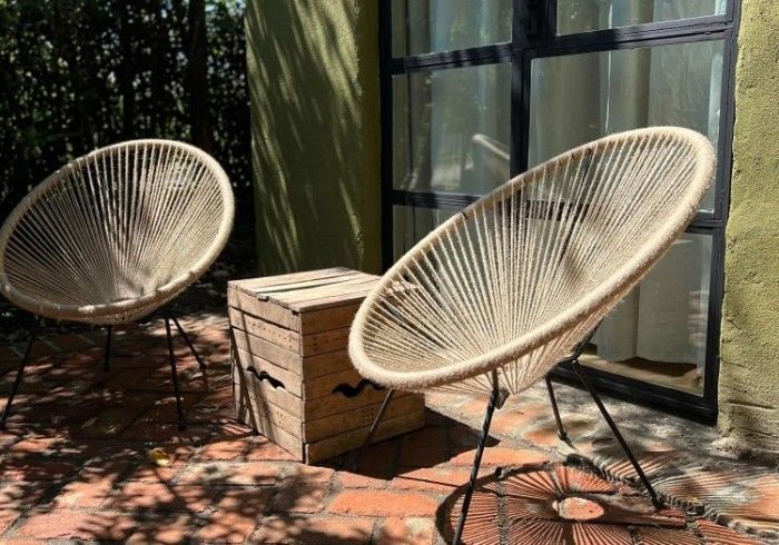 Two beige Acapulco chairs and a wooden crate stool sit on a brick patio in front of a window.