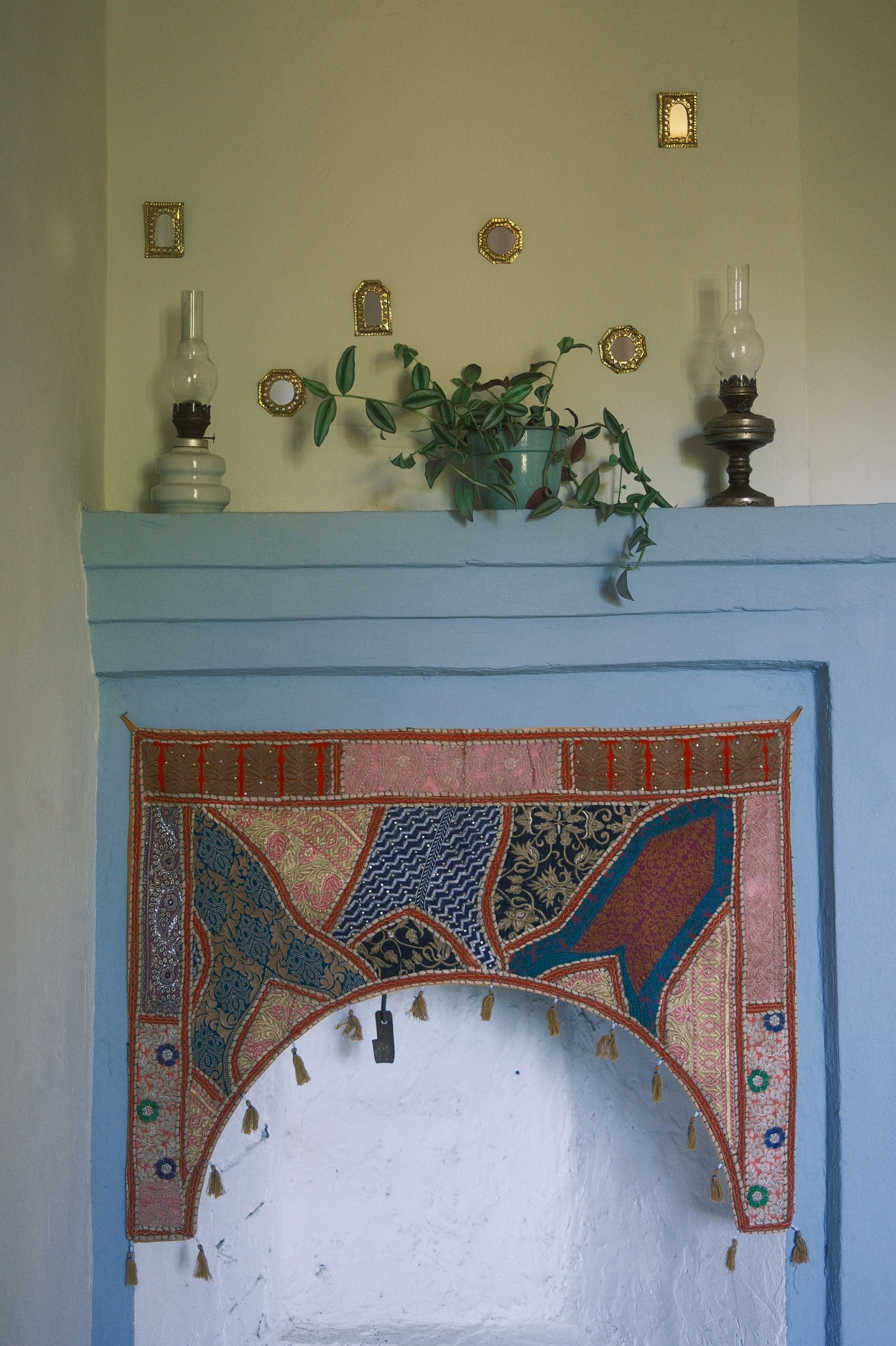 A blue-framed alcove decorated with a colorful, patterned tapestry hanging, topped by a shelf with lamps and a plant.