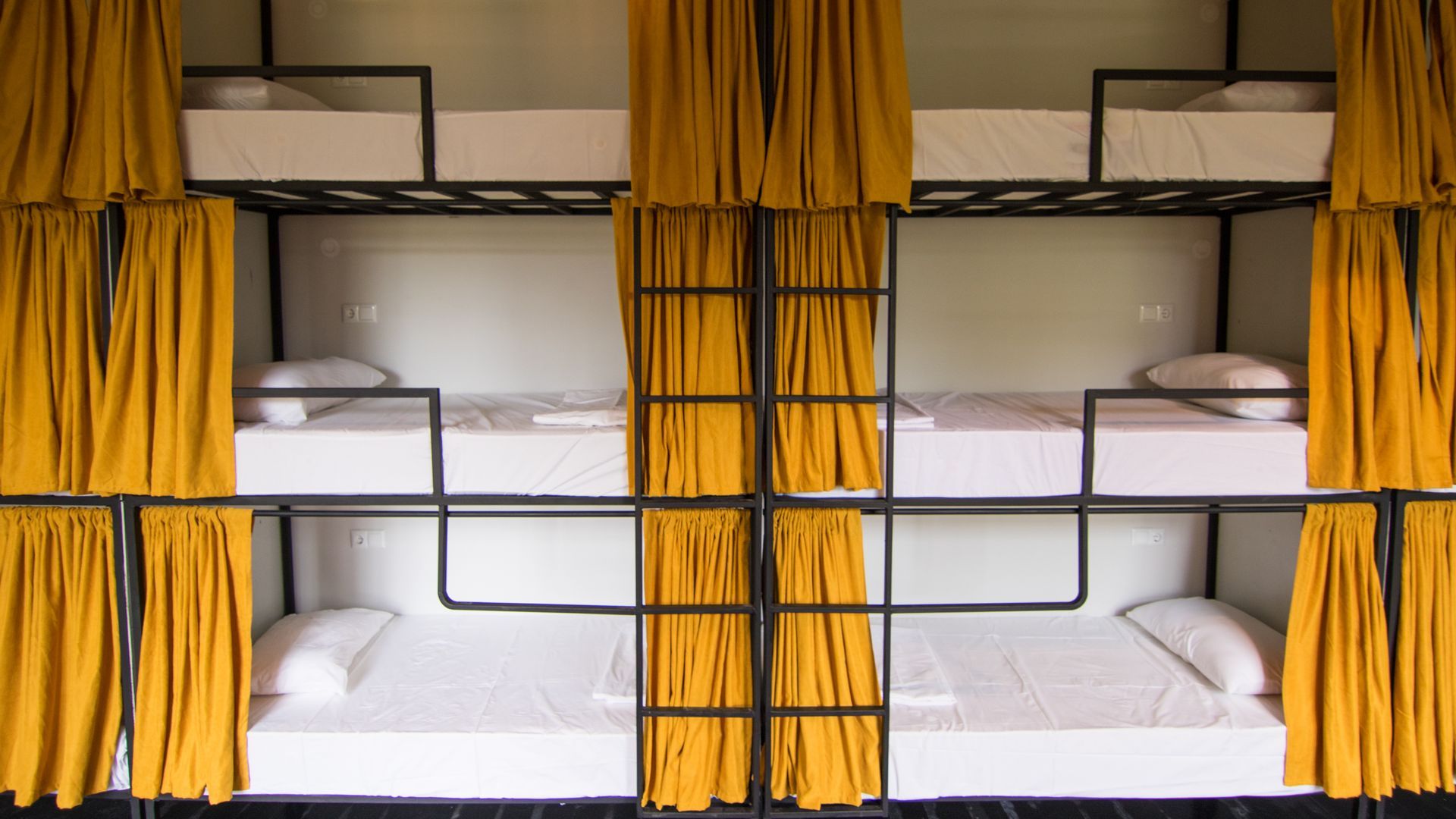 A wall of triple-stacked bunk beds with white linens and mustard yellow privacy curtains in a dormitory setting.